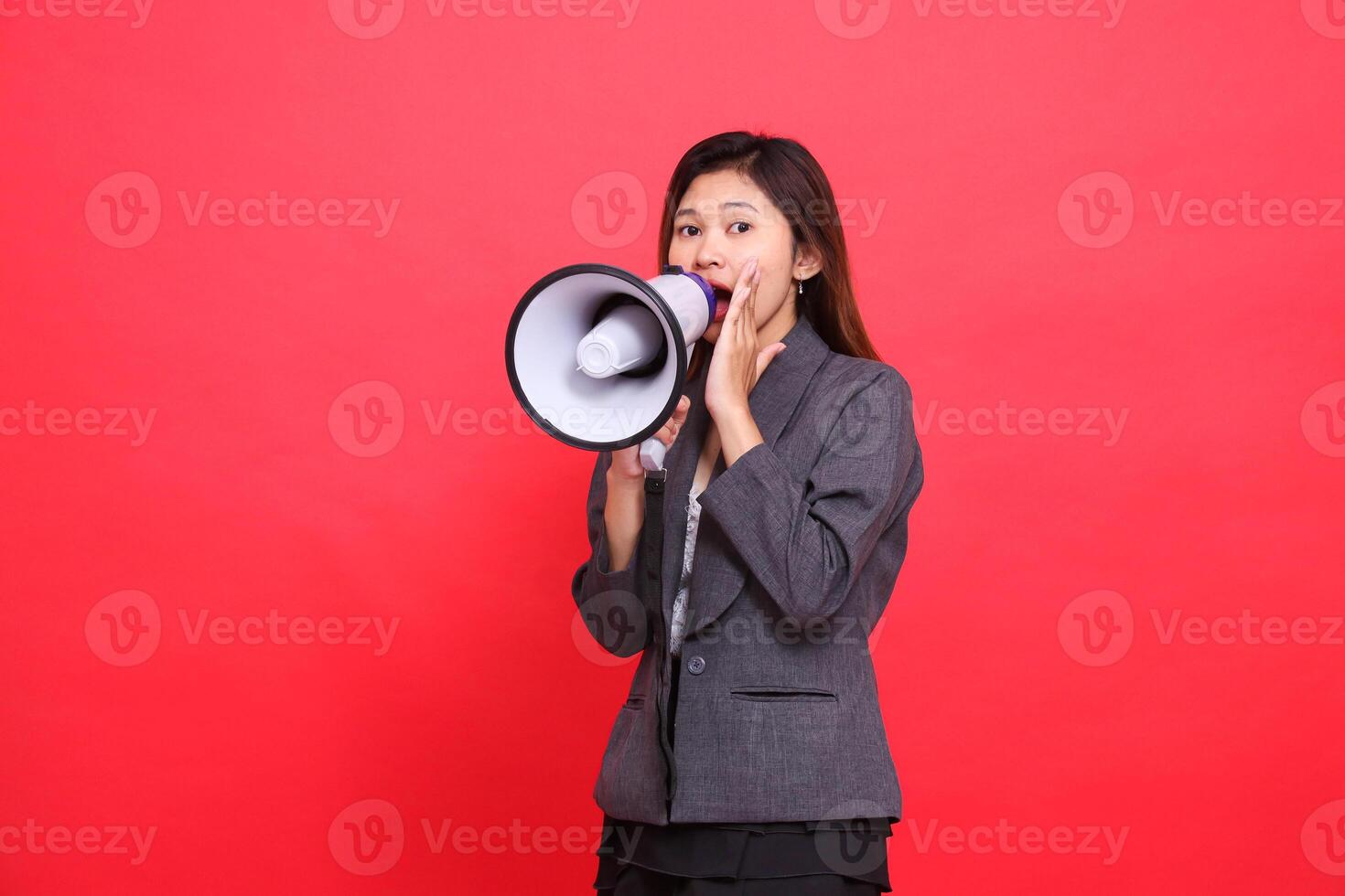 Asian office woman's expression tilted to the right, shouting angrily using a megaphone, hand focusing her voice, wearing a gray jacket and skirt on a red background. for broadcast and advertising photo
