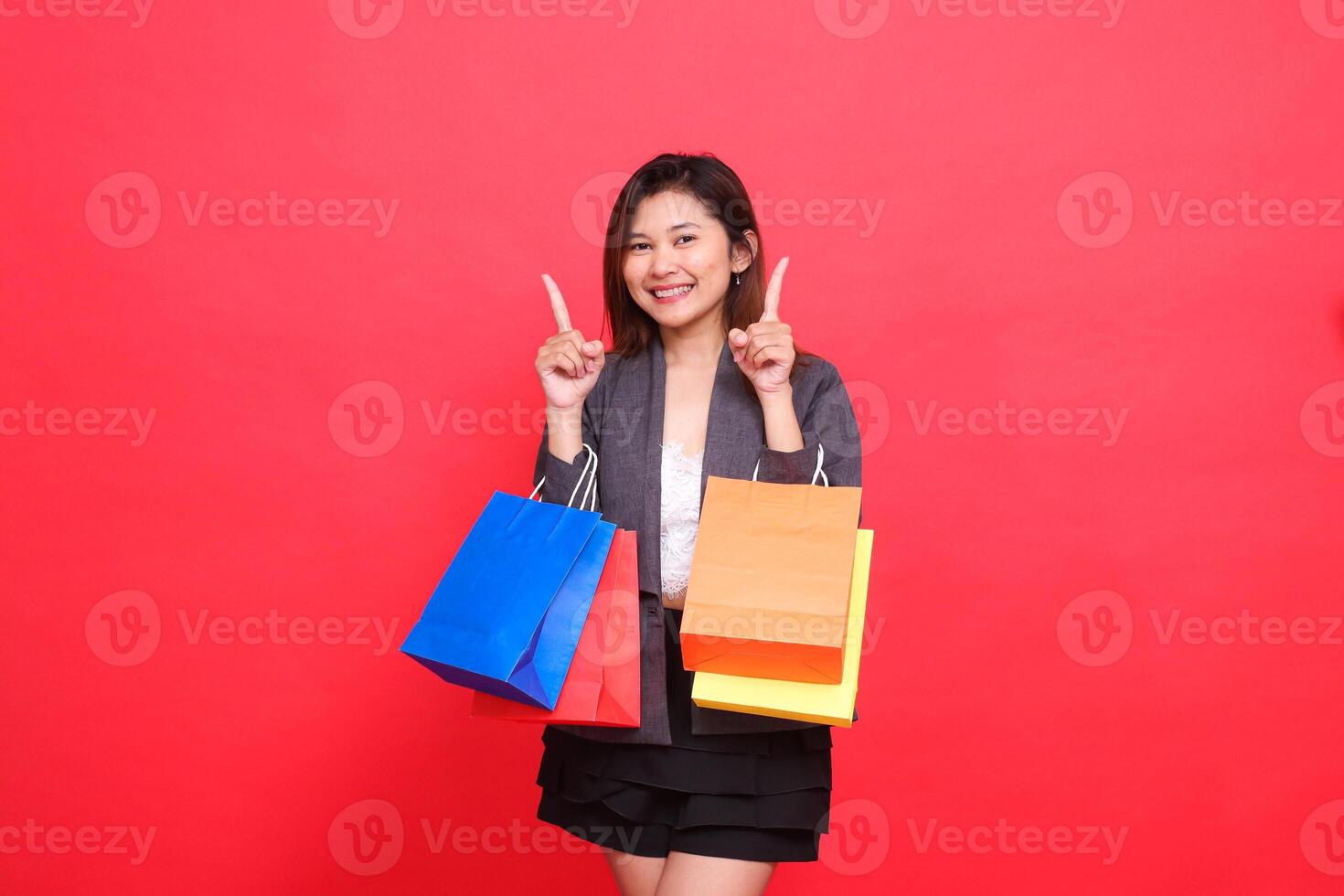 the expression of a cheerful Asian office girl carrying a shopping paper bag with both hands pointing upwards wearing a formal jacket on a red background. for commercial, lifestyle and sales concepts photo