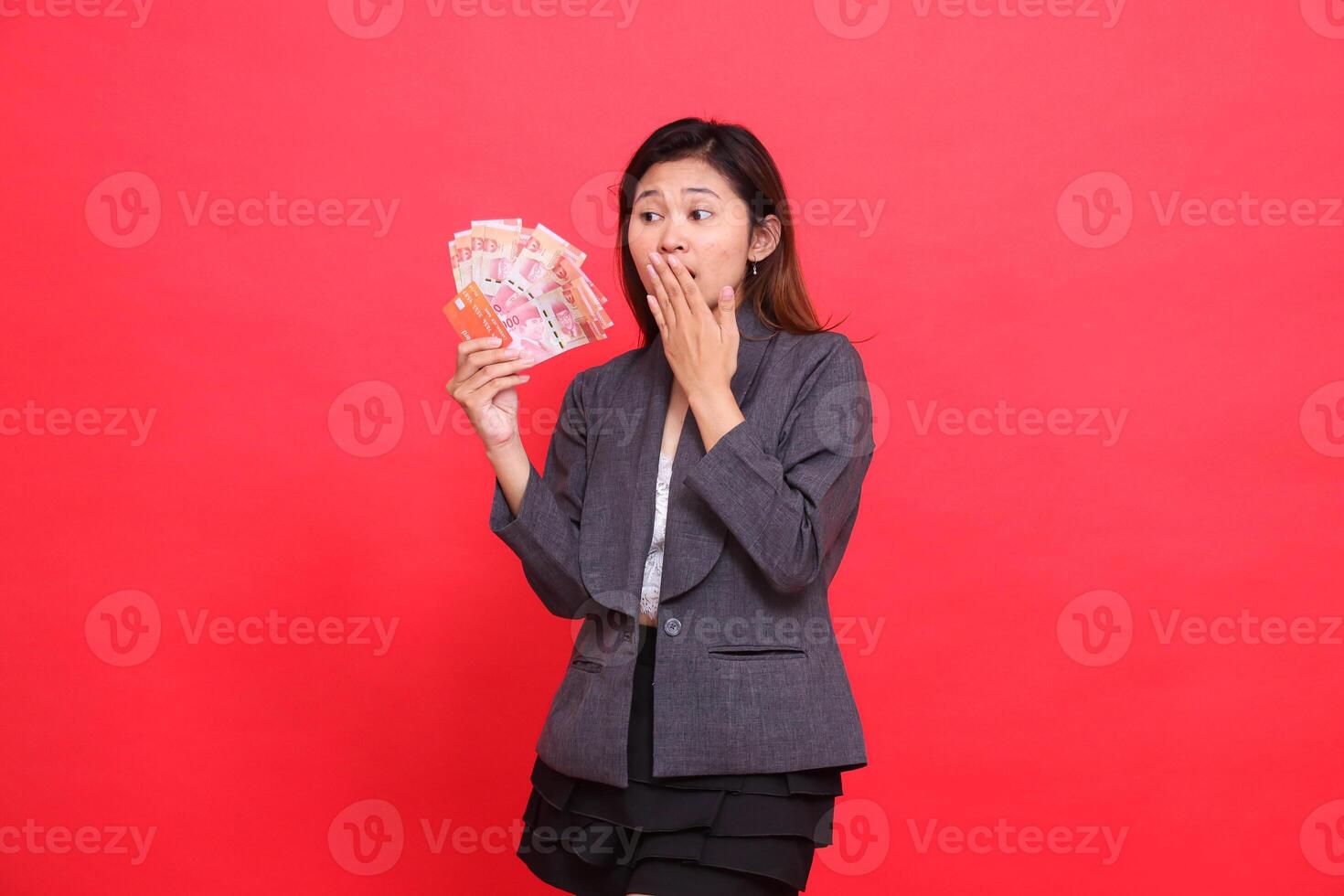 indonesia female director's expression shocked, covering her mouth, hand holding credit, debit and money cards, candid looking at the camera, wearing a gray jacket and skirt with a red background photo