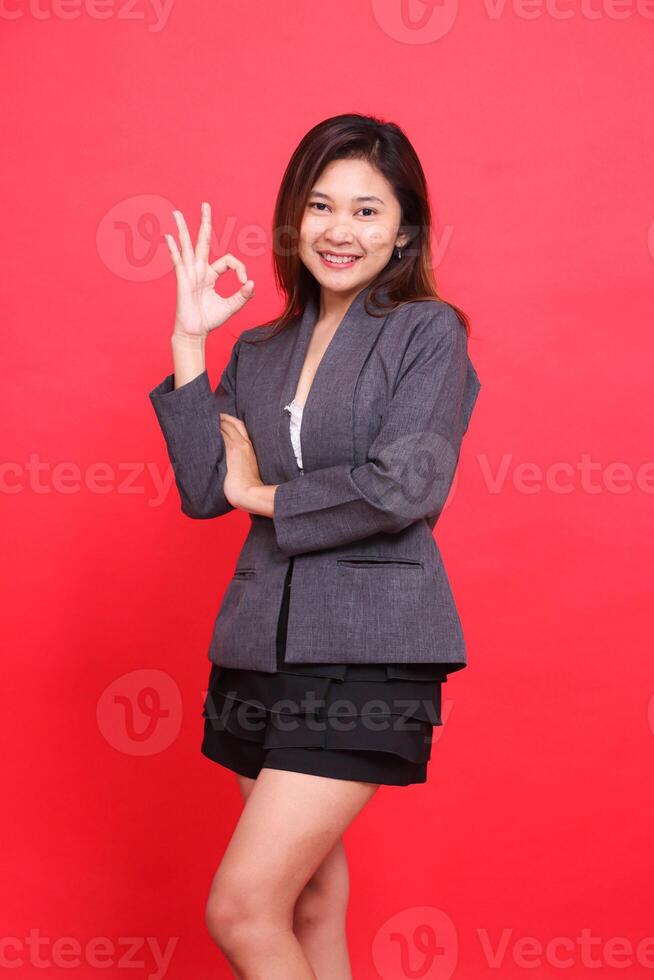 Portrait of a cheerful Asian office girl with her right arm crossed with her arms crossed as an OK sign wearing a gray jacket and black mini skirt on a red background. for corporate concepts photo