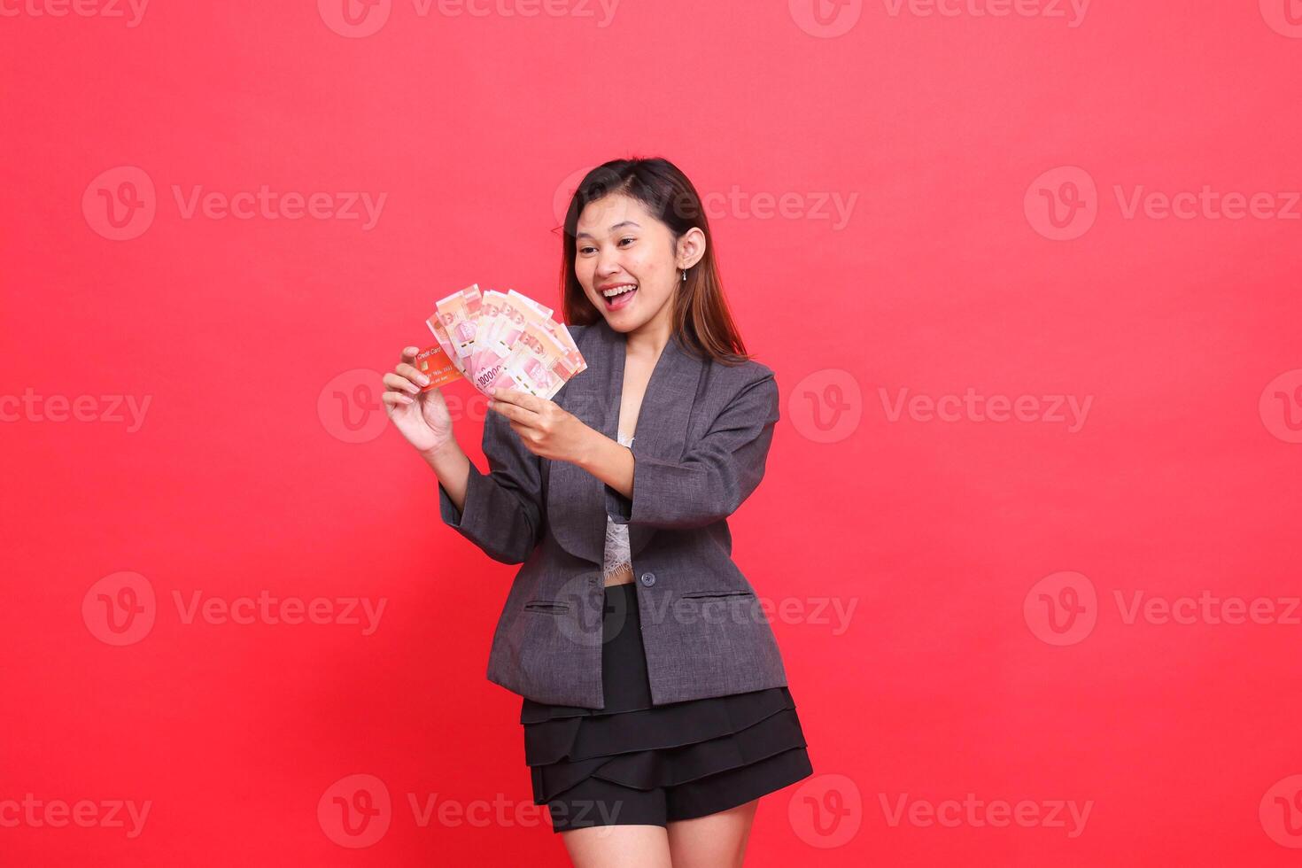 gesture of chinese office woman, candid smile, both hands holding credit cards and money, wearing a jacket and skirt on a red background. for financial, business and advertising concepts photo