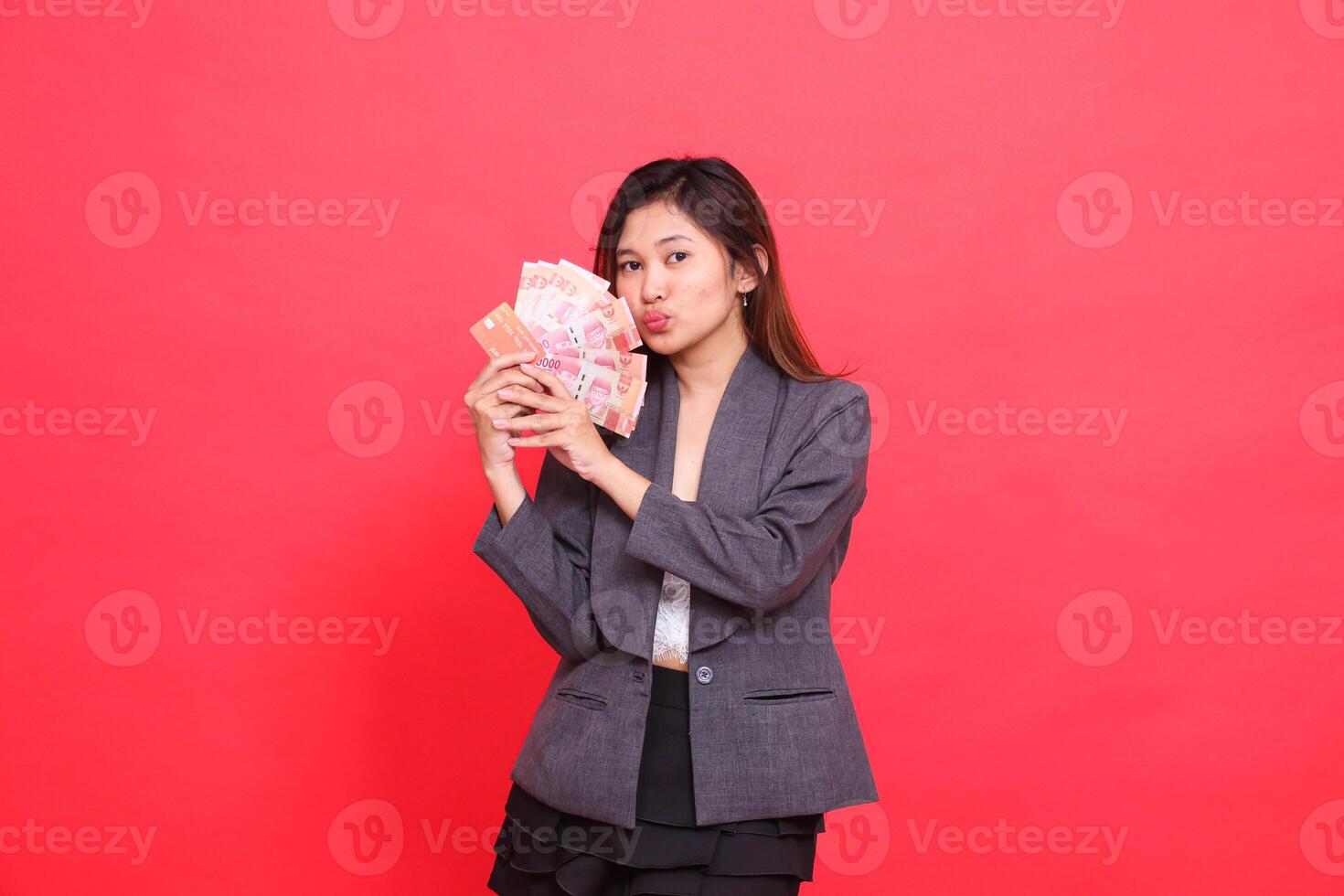 The expression of a cheerful Asian girl director with both hands holding a credit card, debit card and money kissing the camera wearing a gray jacket and skirt on a red background. for advertising photo