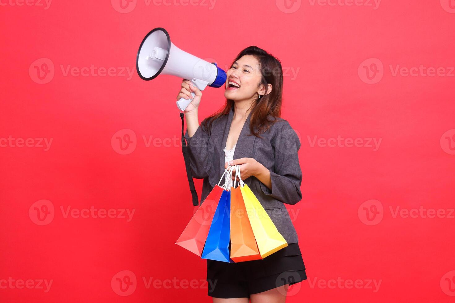 Asian businesswoman shouts cheerfully candidly through a megaphone speaker and carries a shopping paper bag wearing a gray jacket on a red background. for transaction, lifestyle and business concepts photo
