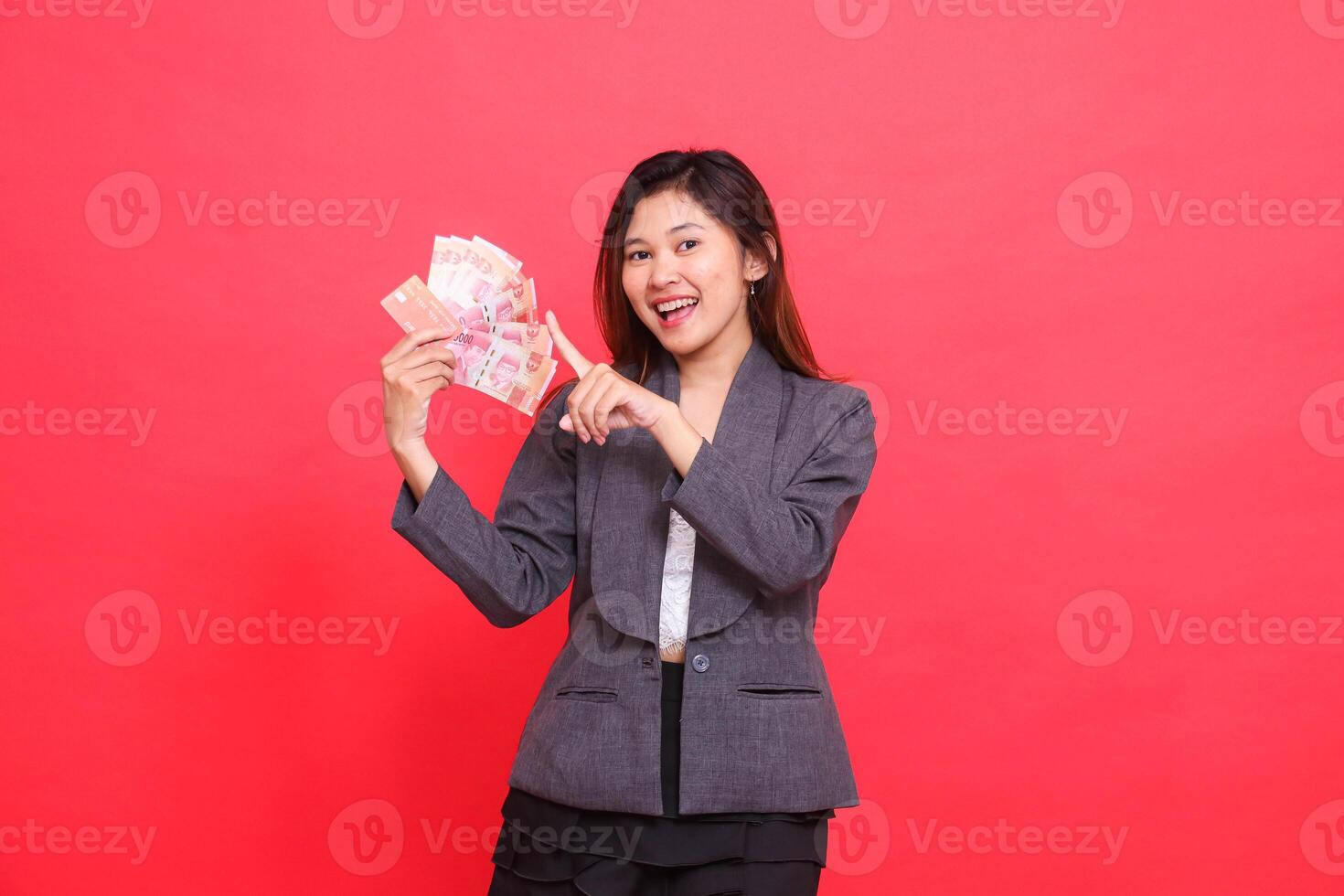 Asian office woman smiling expression holding credit, debit card and money while pointing top right at the camera wearing gray jacket and red skirt. for transaction, business and advertising concepts photo
