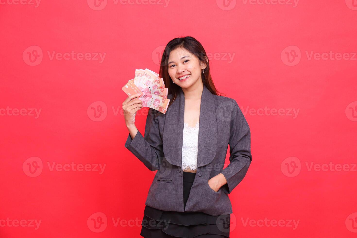 Expression of cheerful indonesia office woman holding debit credit card and money upright and in pocket looking at camera wearing gray jacket and red skirt. for transaction, business and advertising photo