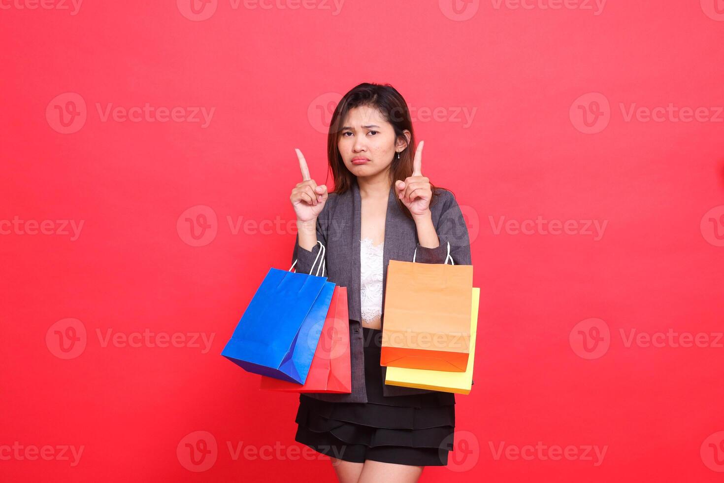 the expression of a sad Asian office woman carrying shopping paper bags with both hands pointing upwards wearing a formal jacket on a red background. for commercial, lifestyle and sales concepts photo