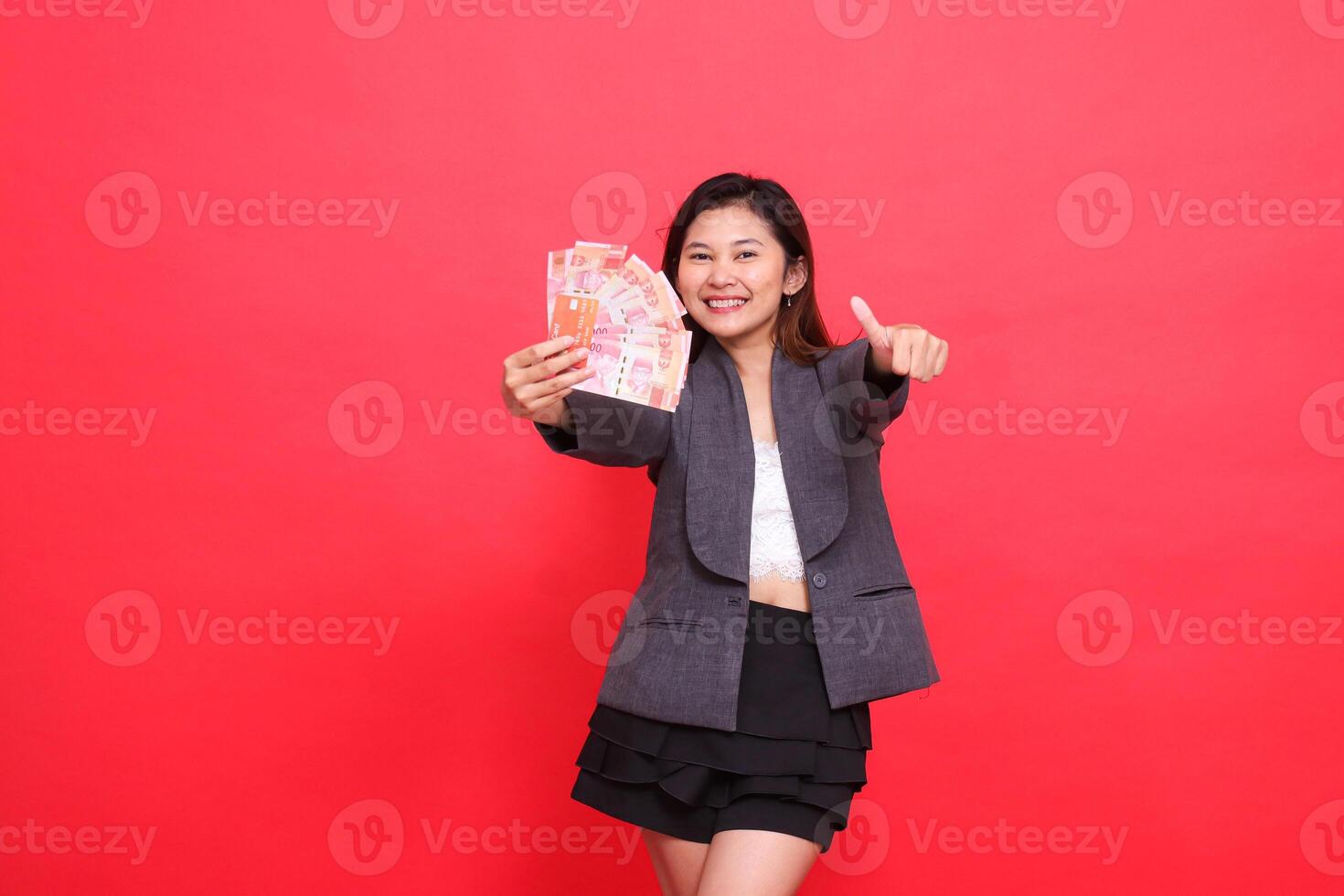 gesture of a happy indonesia career woman holding a credit card and money in front of a thumbs up sign wearing a jacket and skirt on a red background. for transaction, business and advertising photo