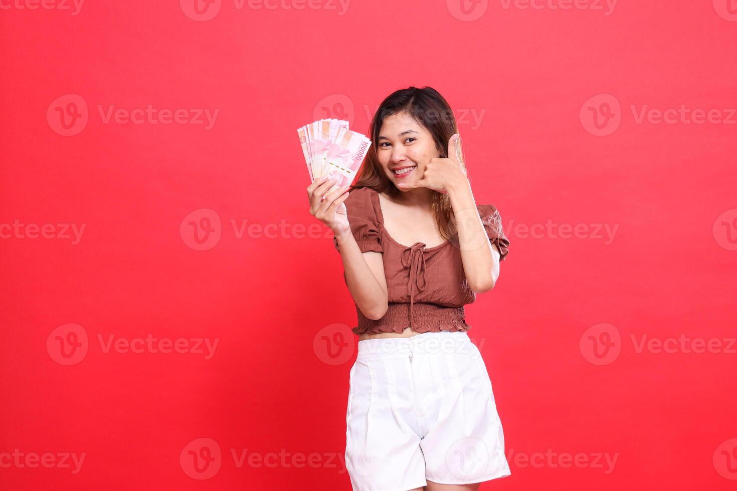 gesture of a happy young indonesia woman holding rupiah money in front and a telephone sign. I am wearing a brown blouse on a red background. for transaction, technology and advertising concepts photo