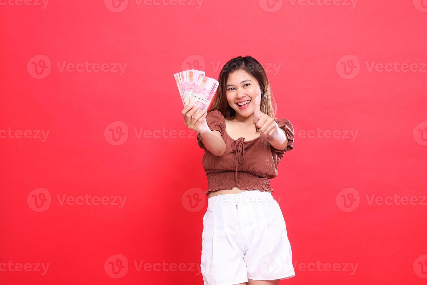 gesture of a cheerful young Asian woman holding rupiah money forward and giving a thumbs up sign okay cool wearing a brown blouse top with a red background. for transaction and advertising photo