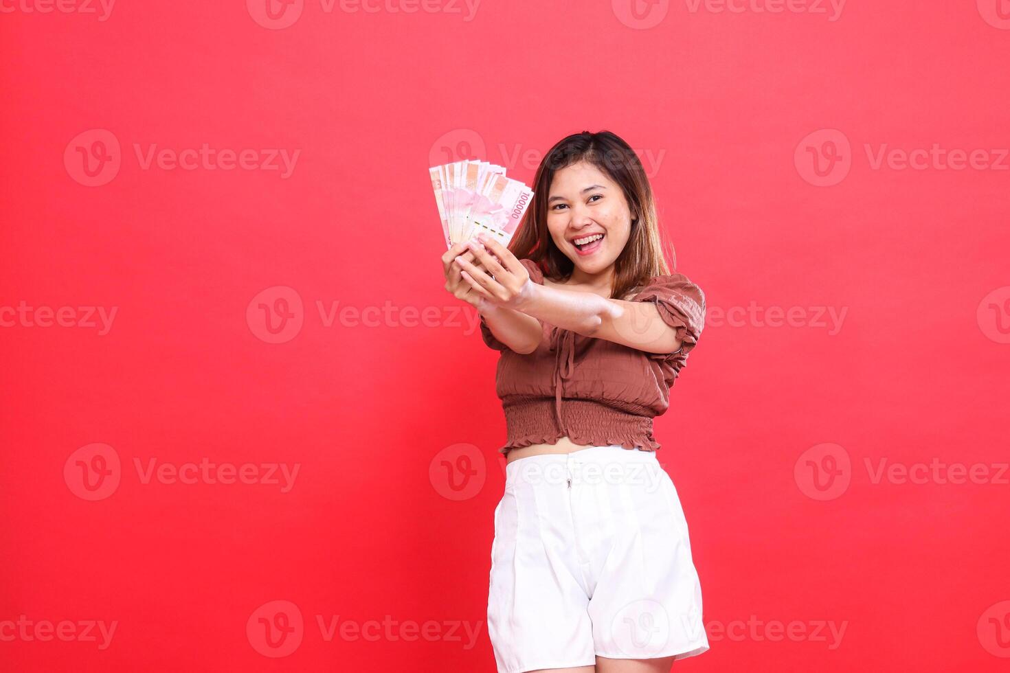 gesture of a cheerful young Asian woman with both hands holding rupiah money in front of her wearing a brown blouse with a red background. for transaction, technology and advertising concepts photo
