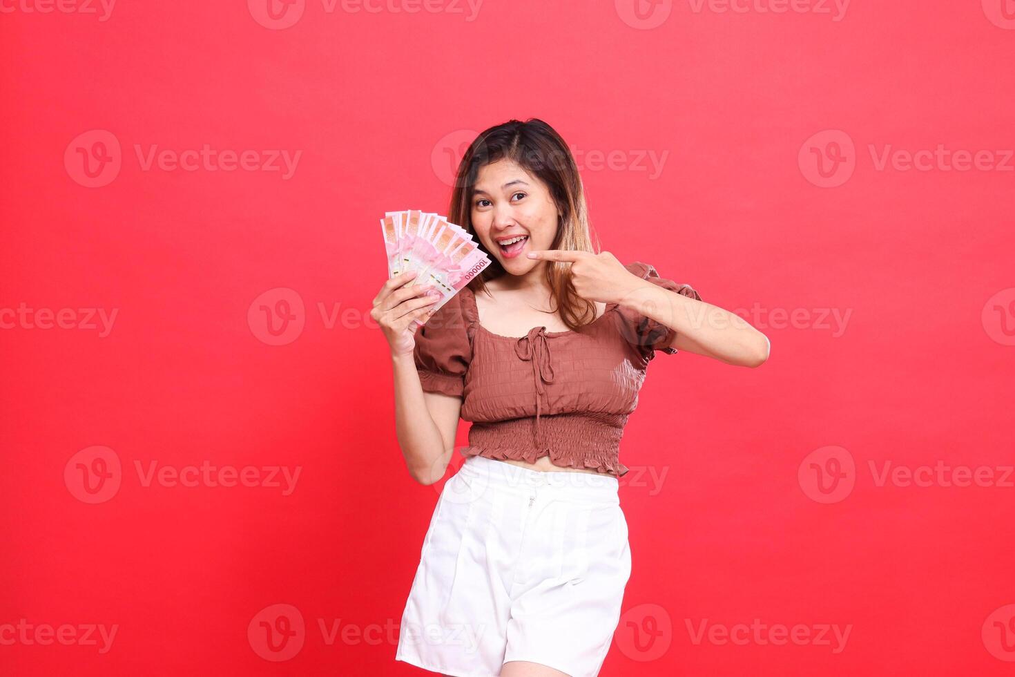 the expression of a cheerful, happy Asian woman holding rupiah money while pointing at it, wearing a brown blouse on a red background. for transaction, technology and advertising concepts photo