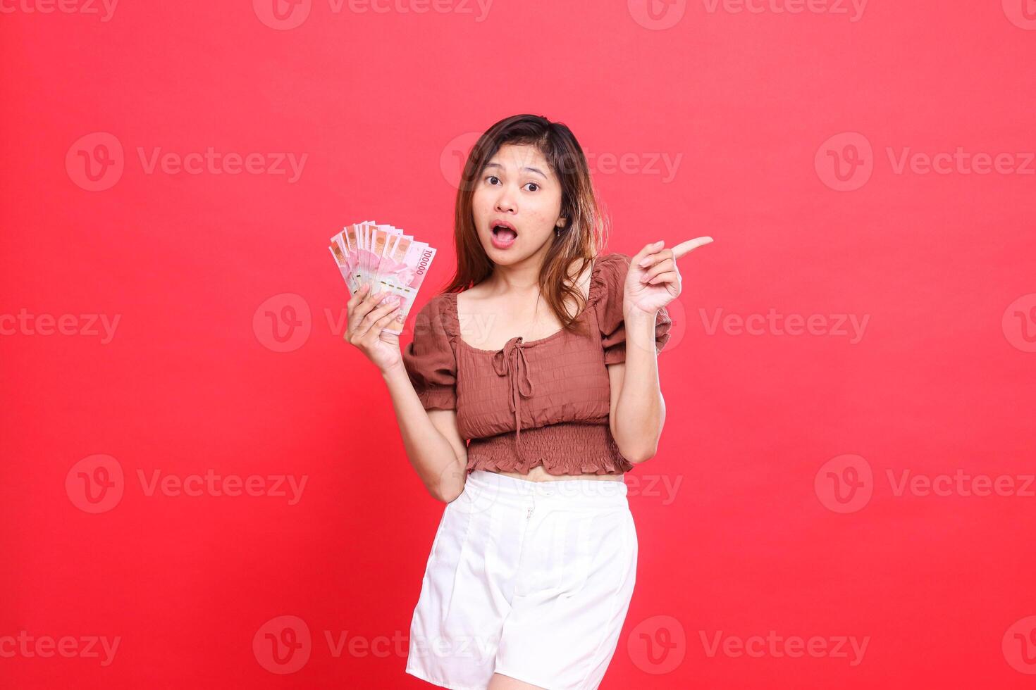The expression of a shocked Asian woman holding rupiah money while pointing to the left wearing a brown blouse with a red background. for transaction, technology and advertising concepts photo