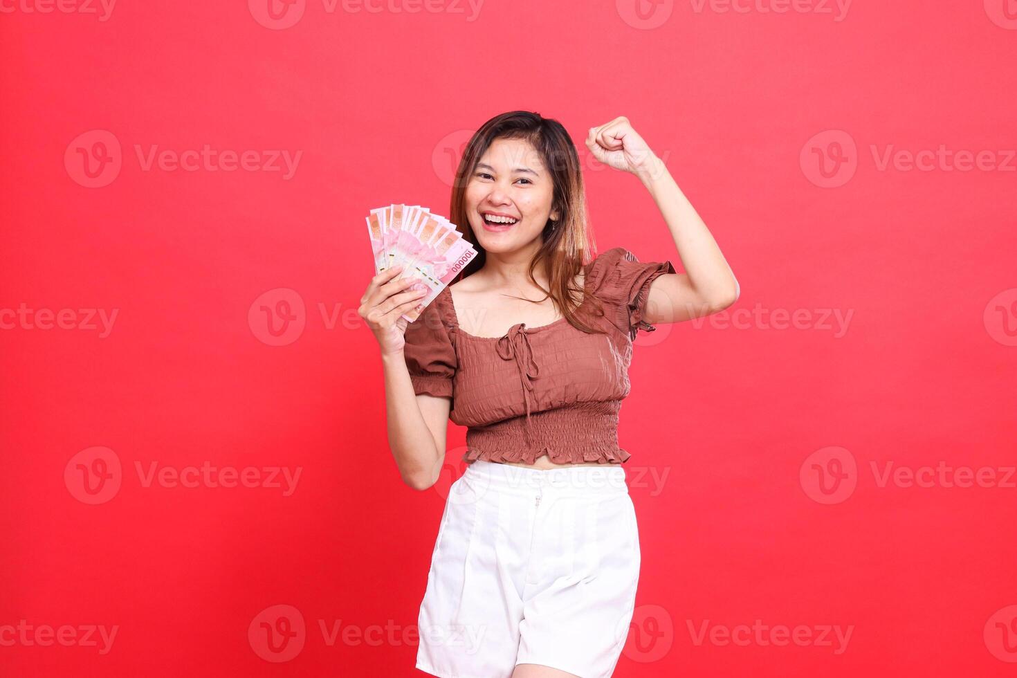 gesture of a cheerful Asian girl clenching her fist upwards successfully while holding rupiah currency wearing a brown blouse top with a red background. for transaction, technology and advertising photo