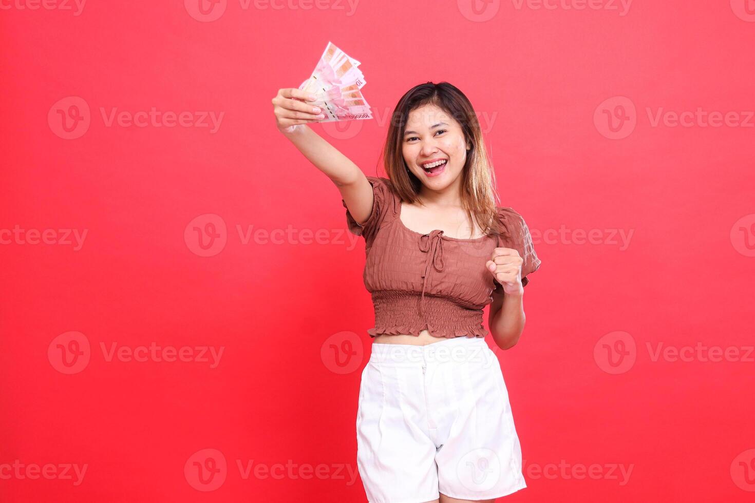 expression of a happy young Asian woman holding rupiah bills up and holding her hands to the side wearing a brown blouse top on a red background. for transaction, technology and advertising concepts photo