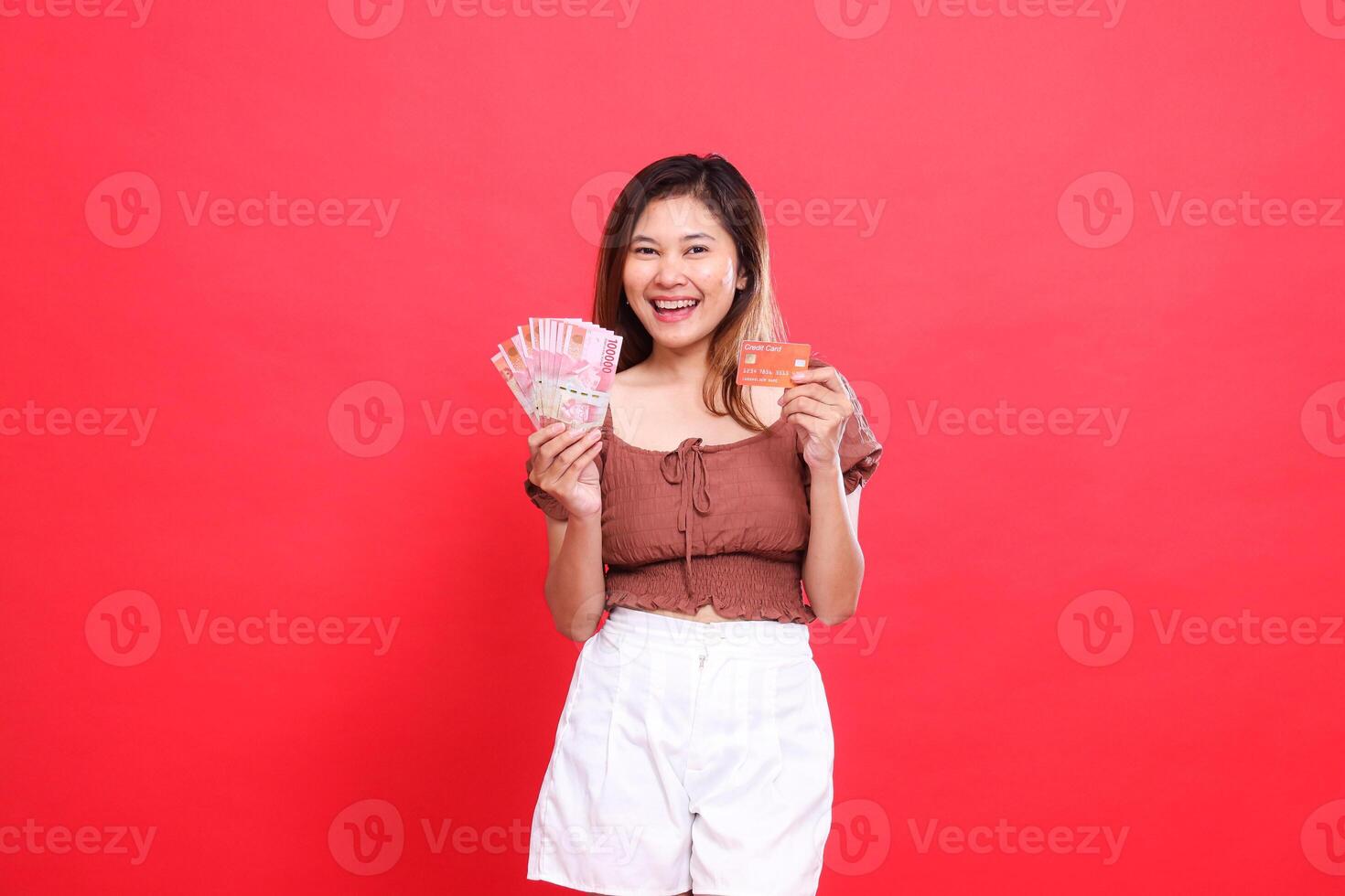 Cheerful indonesia woman holding rupiah money and debit credit card wearing brown blouse top with red background. for transaction, technology and advertising concepts photo