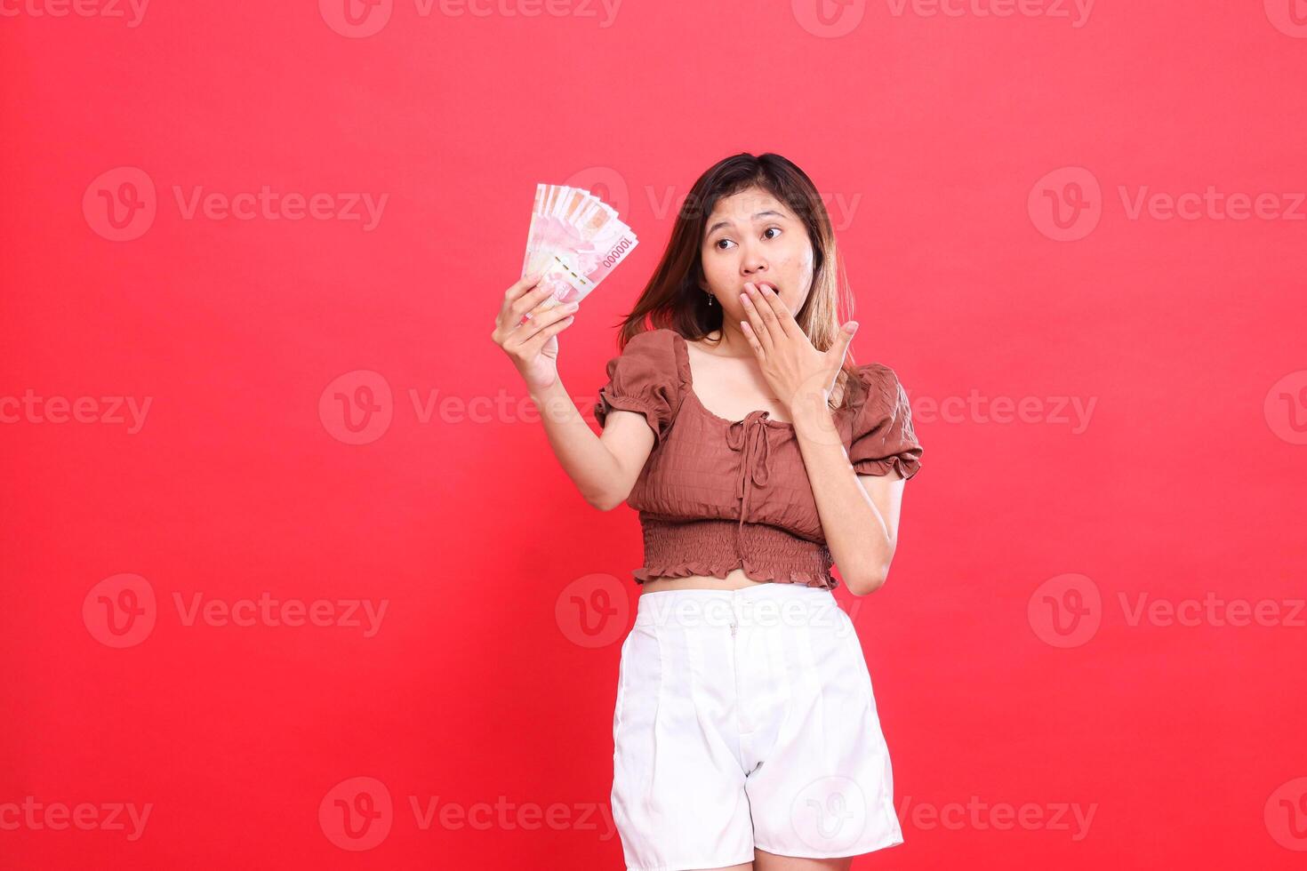 The expression of a young indonesia woman is shocked, covering her mouth and holding rupiah bills up, wearing a brown blouse on a red background. for transaction, technology and advertising concepts photo