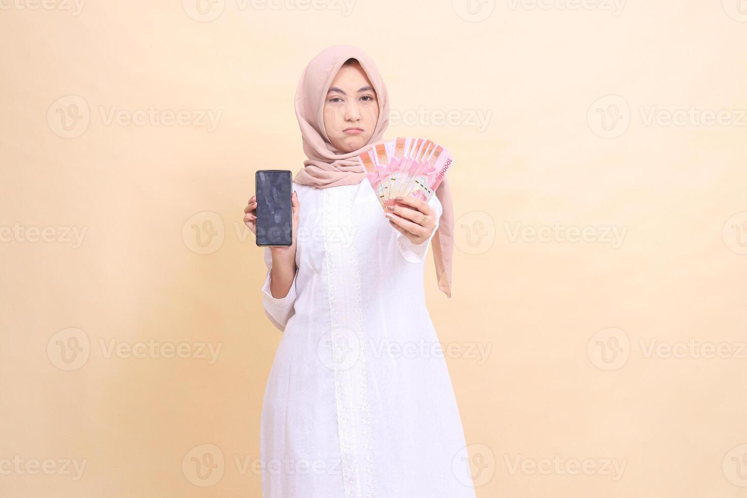 Young Asian Muslim woman wearing a hijab frowns holding a cellphone gadget and rupiah money in her hand. Lifestyle, business and commercial concept photo