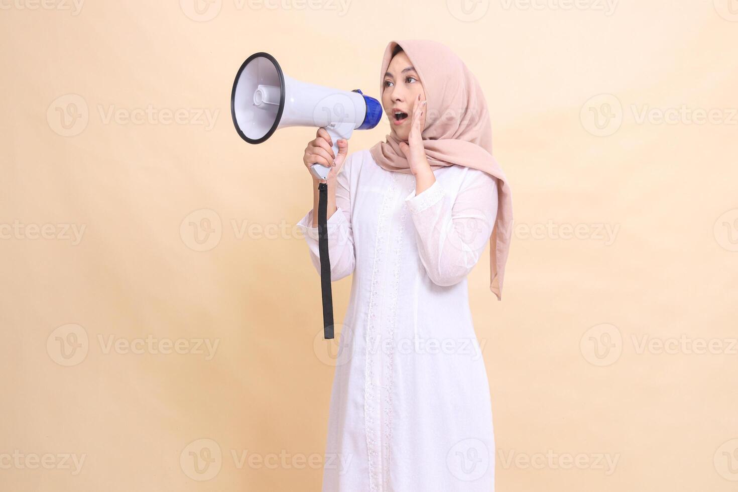 Asian Muslim woman wearing hijab slanted to the right candidly shouting angrily holding a loudspeaker megaphone while focusing her voice. Technology, broadcast and promotion concept photo