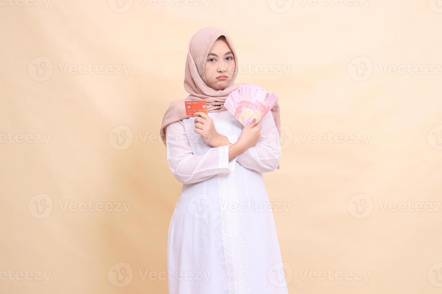 woman muslim indonesia in hijab frowning holding rupiah cash and credit card in crossed hands. Lifestyle, religious, business and finance concepts photo