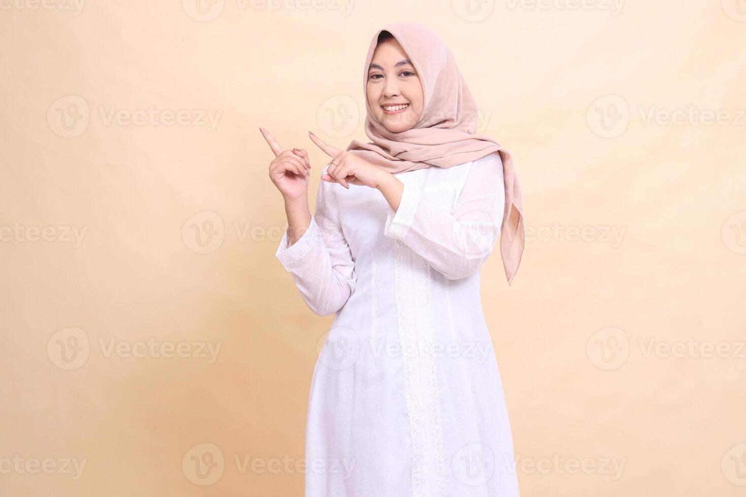 young asian muslim woman standing to the camera smiling cheerfully with arms crossed to the top right. Fashion, pointer, business and promotion concept photo