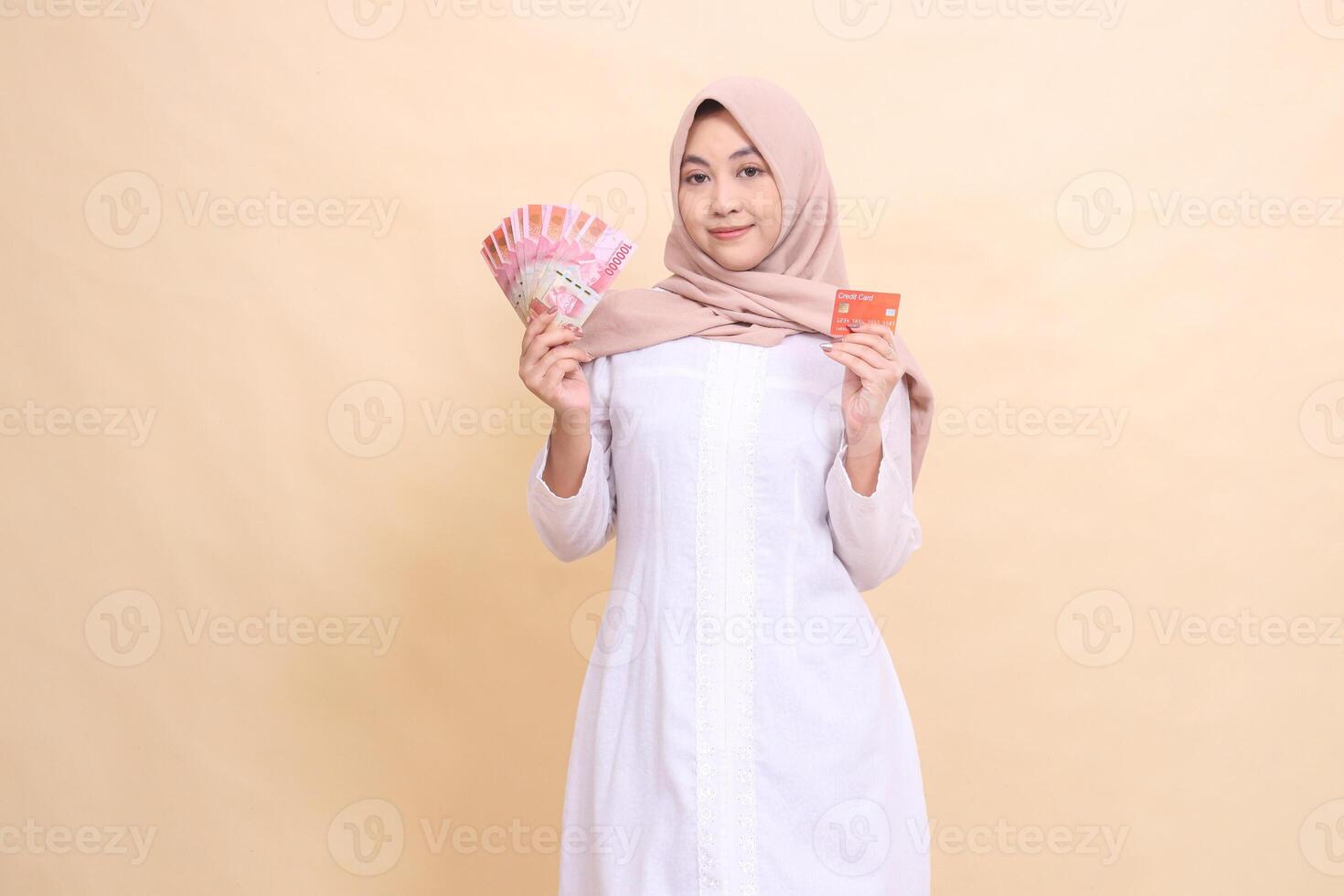 Asian Muslim woman in hijab smiles holding rupiah cash and credit card in hand. Lifestyle, religious, business and finance concepts photo