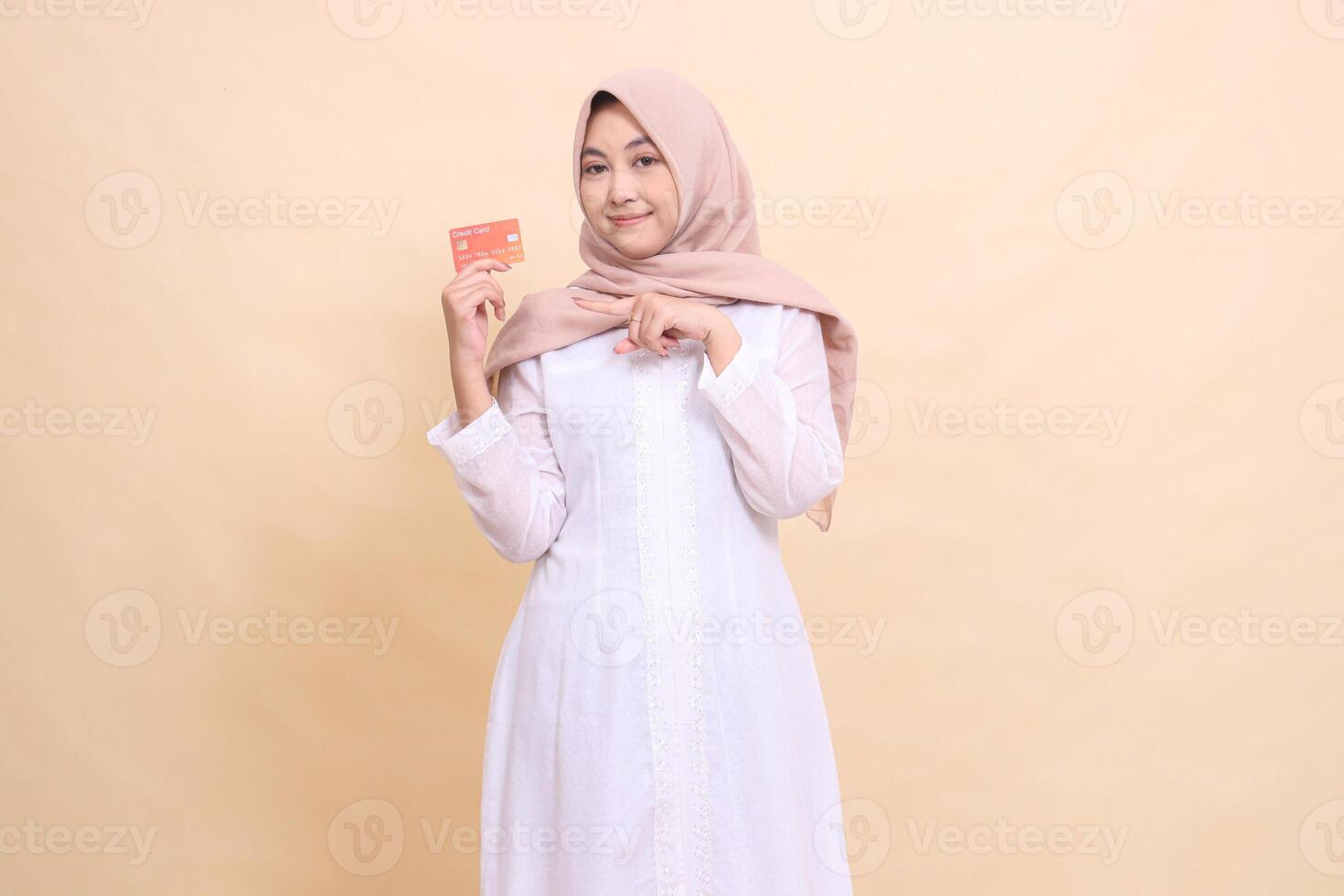 asian muslim woman in hijab smiling holding debit credit card in hand while pointing at it. Lifestyle, business and finance concept photo