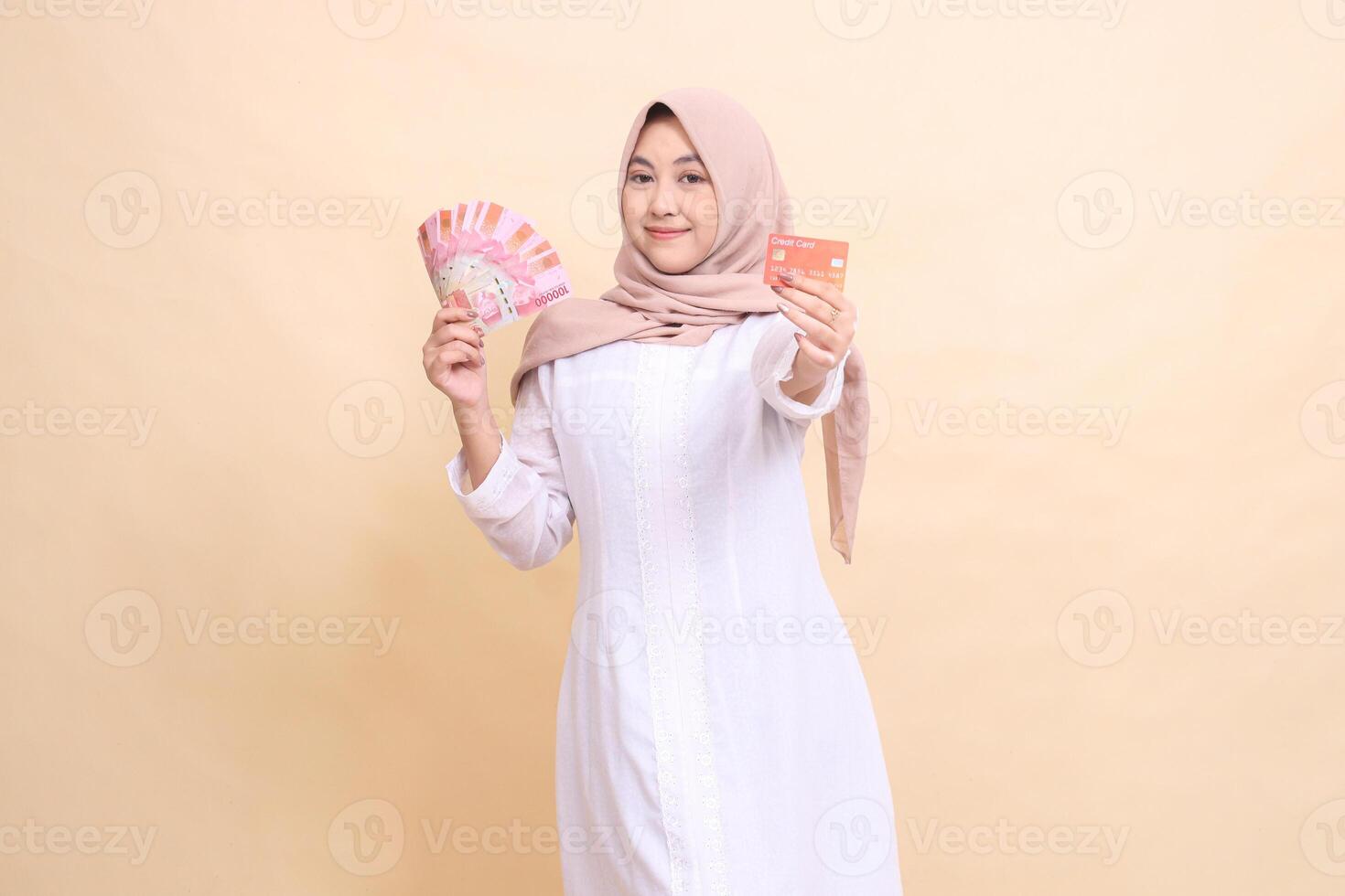Asian Muslim woman wearing a hijab smiles gracefully holding rupiah cash and credit cards in her hands. Lifestyle, religious, business and finance concepts photo