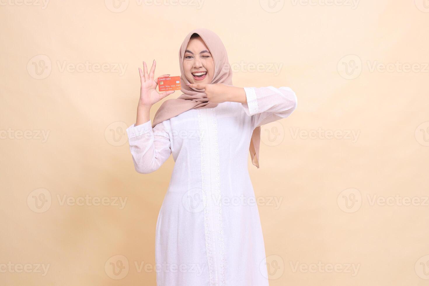 Asian Muslim woman wearing a hijab smiling cheerfully, winking, holding a debit credit card in her hand and pointing up. Lifestyle, business and finance concept photo
