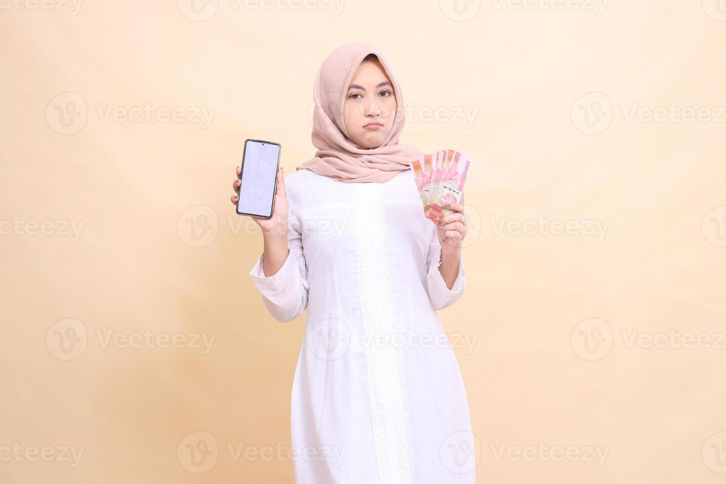 Young Asian Muslim woman wearing a hijab frowns holding a cell phone gadget and rupiah money in her hand. Lifestyle, business and commercial concept photo