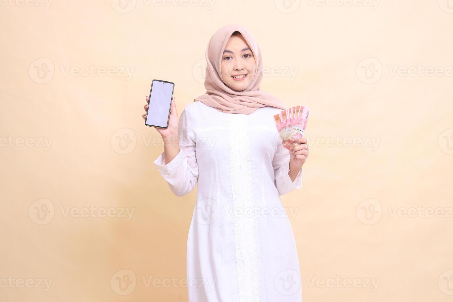 Young Asian Muslim woman wearing a hijab smiles holding a cell phone gadget and rupiah money in her hand. Lifestyle, business and commercial concept photo