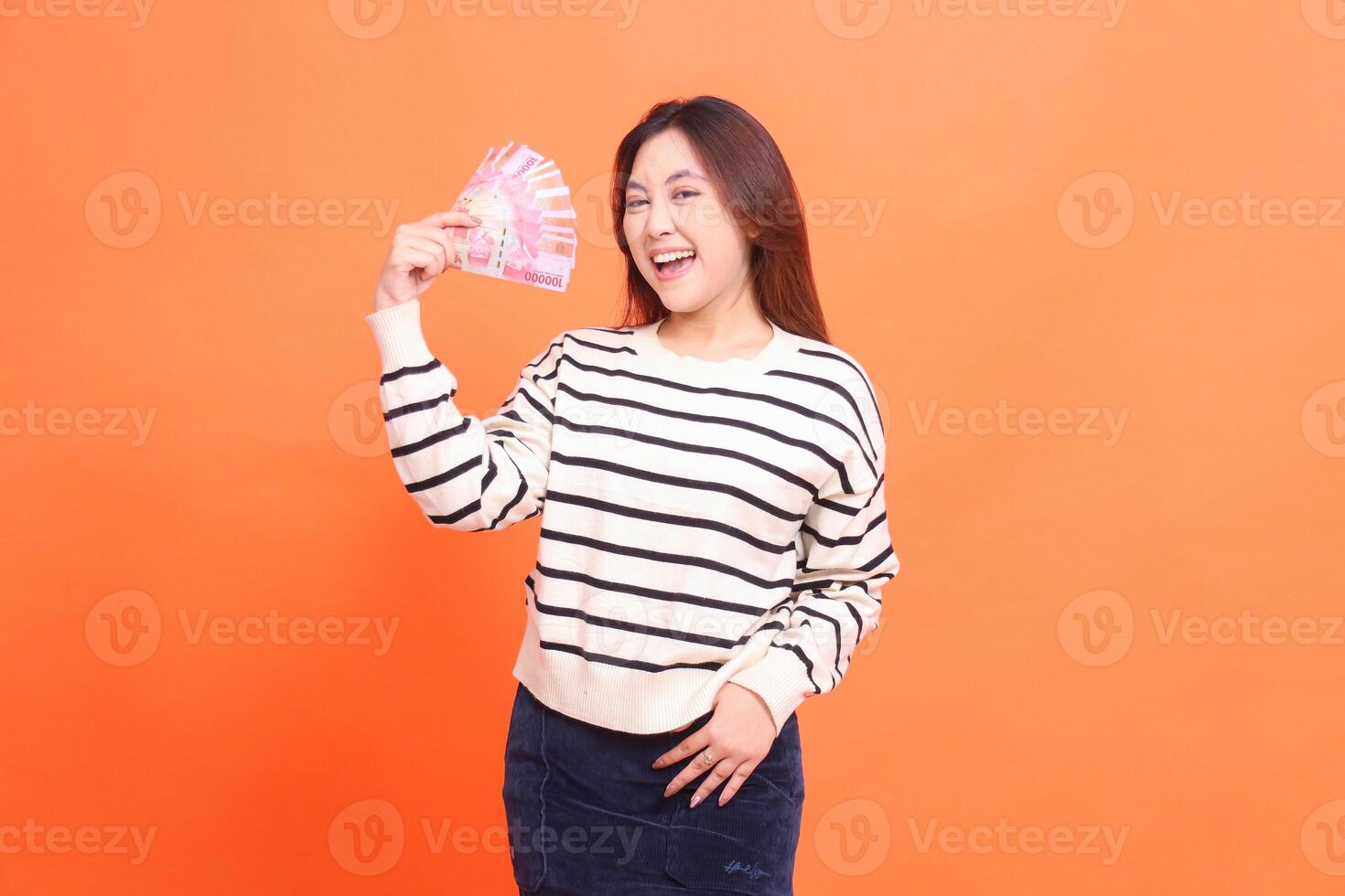 an Asian girl with a cheerful expression at the camera stands while holding several rupiah notes and holding her skirt. for fashion, business and finance concept photo