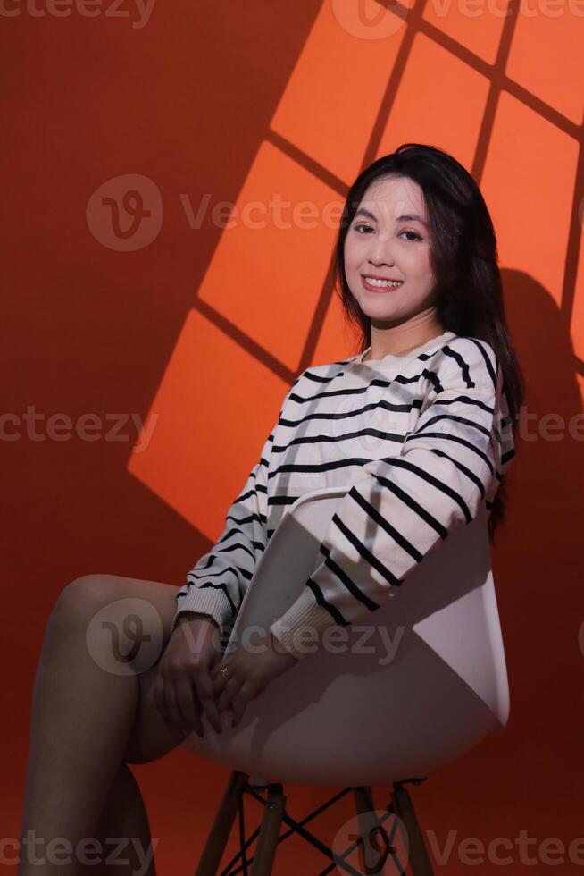 A beautiful smiling asian girl sits on a chair in a dark studio, light shines on her through the curtains, forming a window pattern. Portrait of woman in projection light spotlight in studio photo