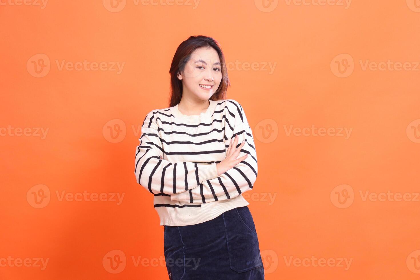 confident cheerful beautiful asian girl standing with arms crossed and looking at camera isolated on orange background wearing white sweater photo