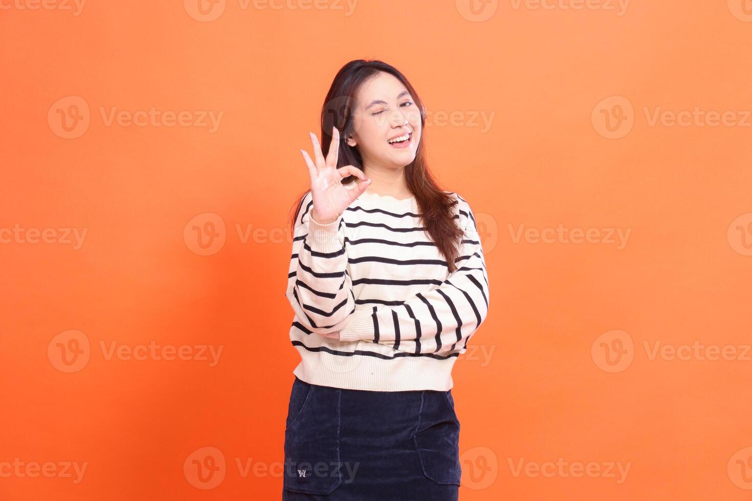 beautiful happy indonesia girl winking confident standing with arms crossed gesturing okay good cool and looking at camera isolated on orange background wearing white sweater photo
