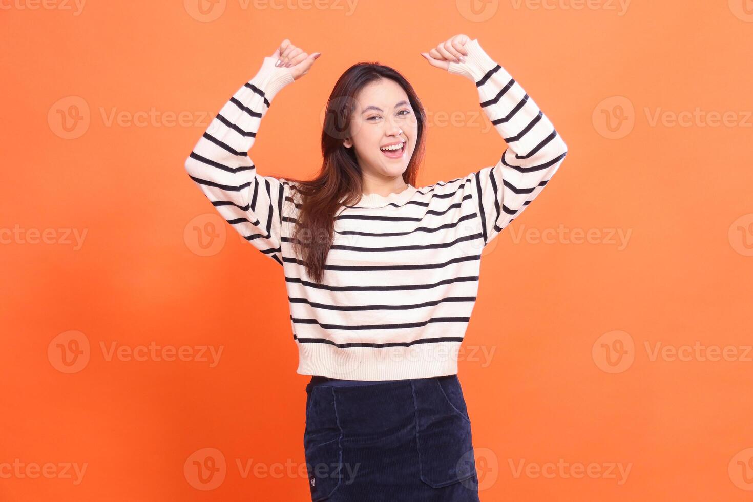 beautiful happy confident asian woman standing with clenched fist up and looking at camera isolated on orange background wearing white sweater photo