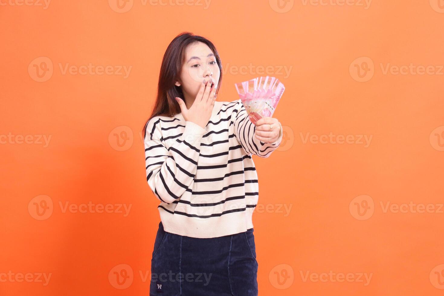 The expression of a shocked Asian girl candidly stands while holding a number of rupiah bills in front of her and covering her mouth. for fashion, business and finance concept photo