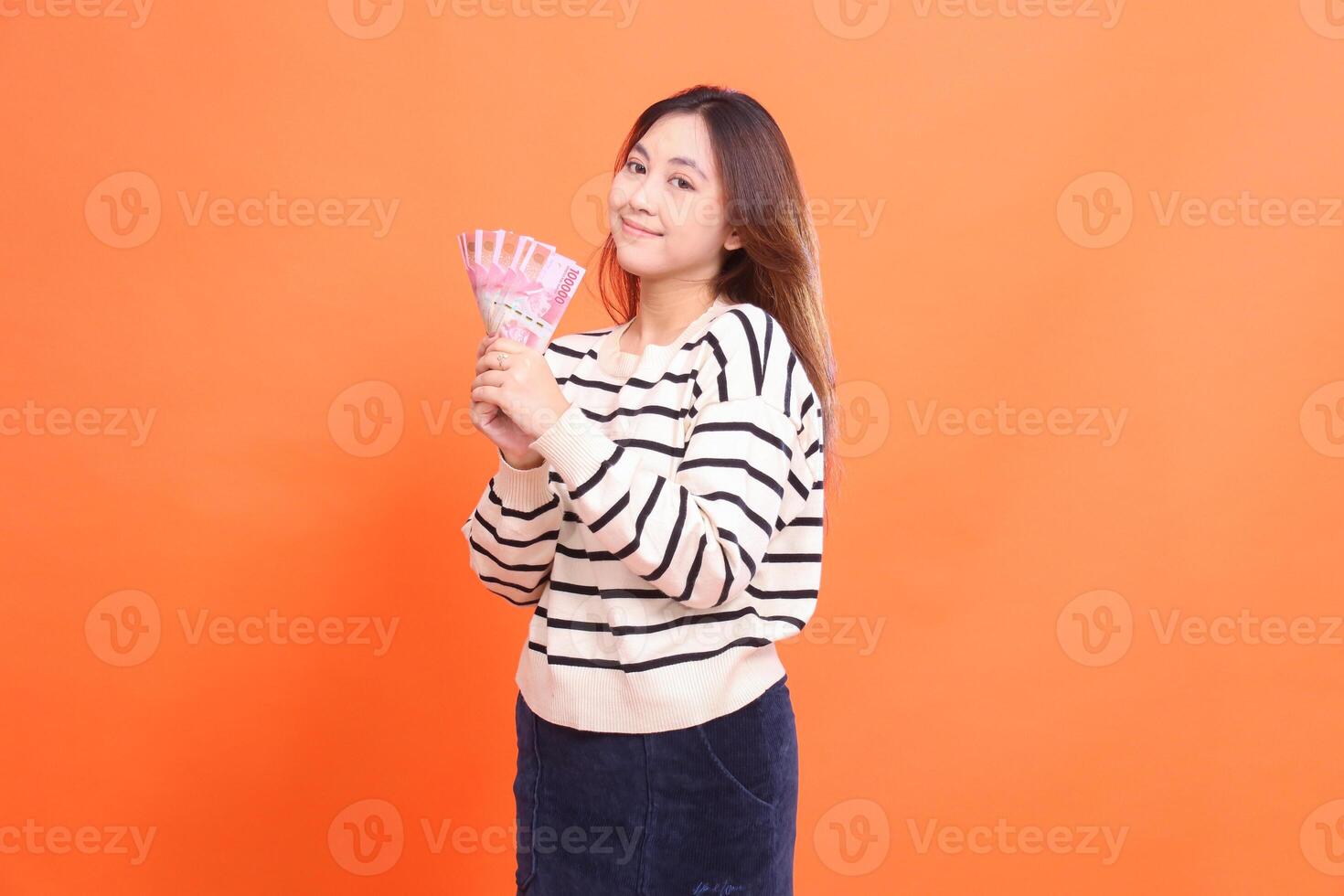 An Asian woman with a smiling expression on her right side stands while holding several rupiah notes in both hands. for fashion, business and finance concept photo