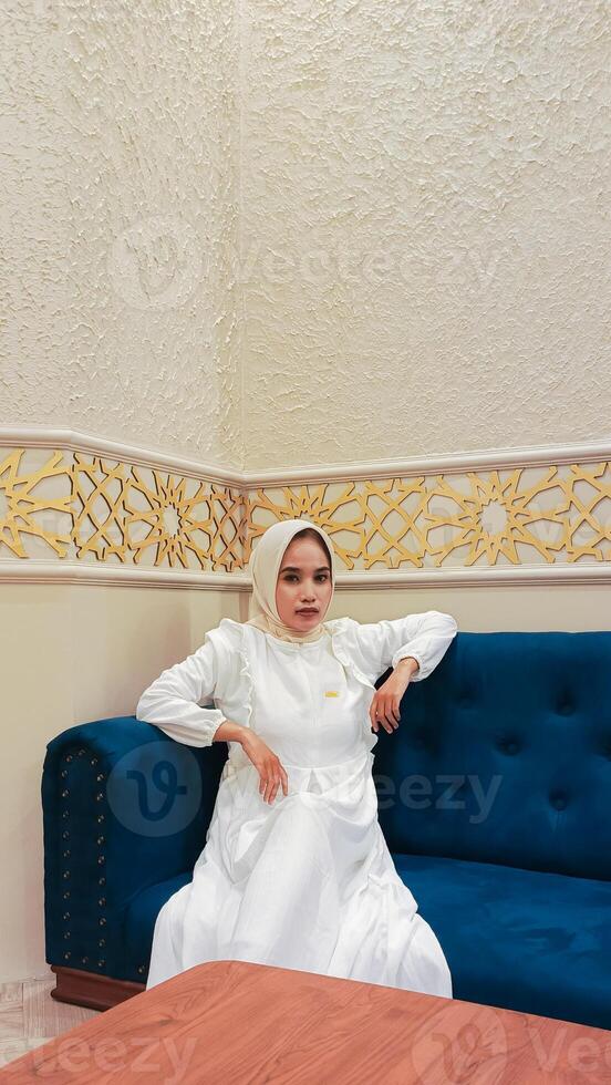 portrait of a beautiful indonesia woman with an elegant expression, both hands resting and sitting on a sofa with an Islamic ornamental calligraphy background, Arabic atmosphere photo