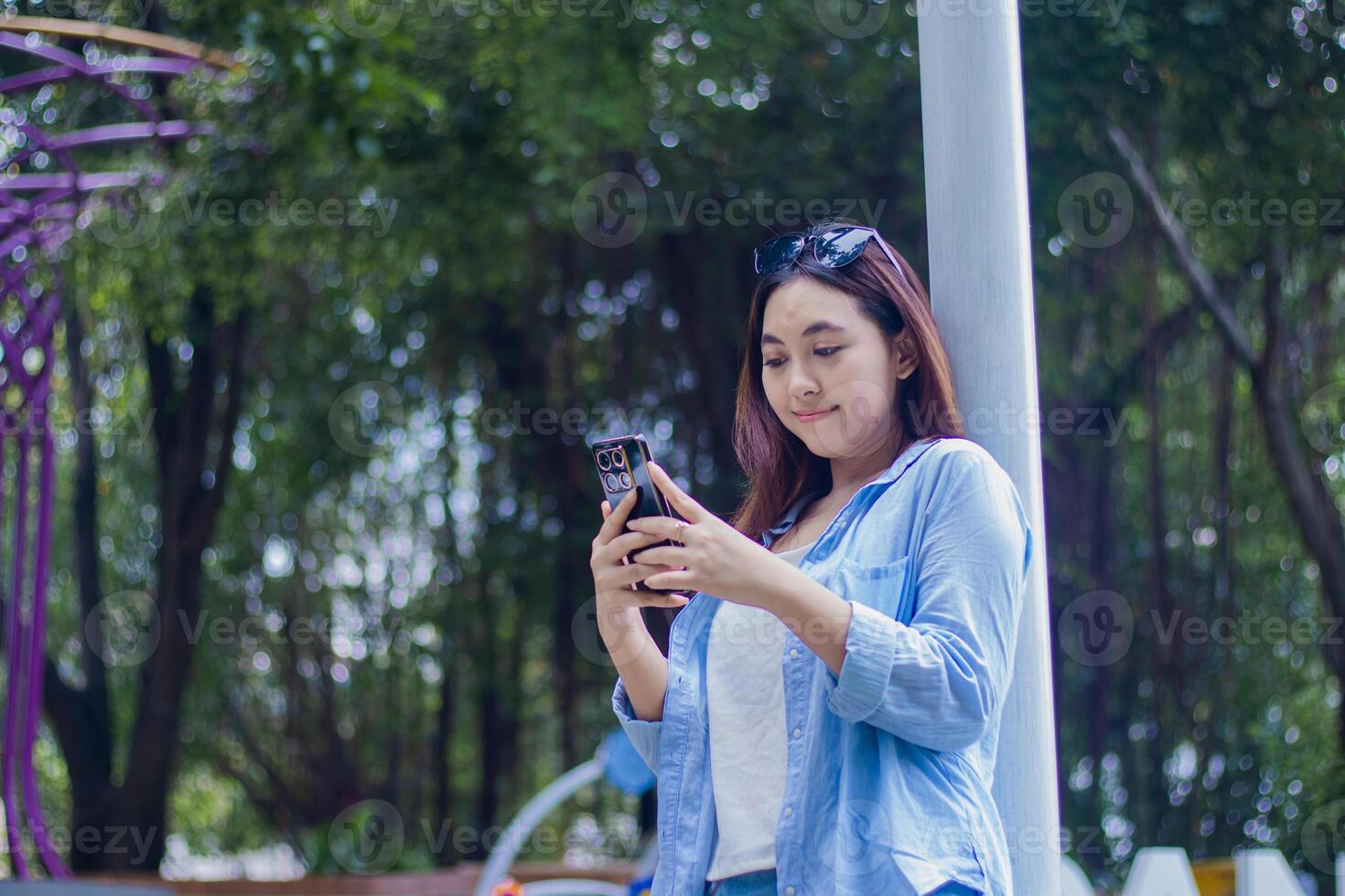Beautiful smiling Asian woman leaning on the fence operating a smartphone gadget in the campus park in sunny weather. female student for education, promotion and lifestyle concept photo