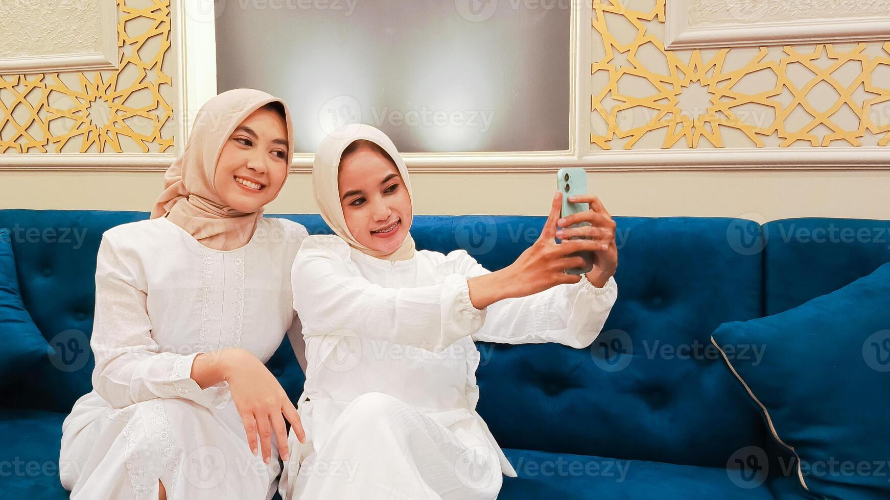 portrait of a pair of beautiful Asian women sitting on the sofa smiling happily taking selfies in front of their cell phones with an elegant Arabic nuance in the background of Islamic calligraphy photo