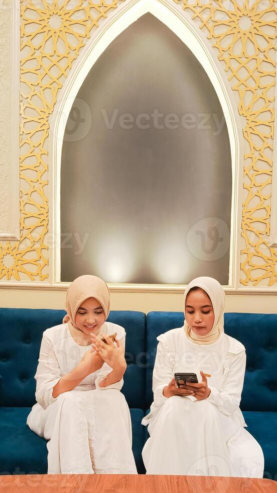 a pair of women sitting on a sofa operating their respective cellphone gadgets with an elegant Arabic Muslim calligraphy background photo