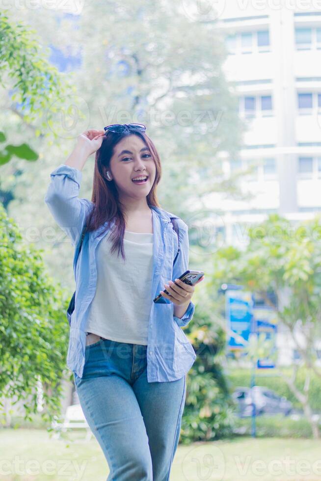 Cheerful beautiful Asian woman looking at the camera using smart phone while wearing earphones at the campus park on a sunny summer day. female students for education, promotion and lifestyle concepts photo