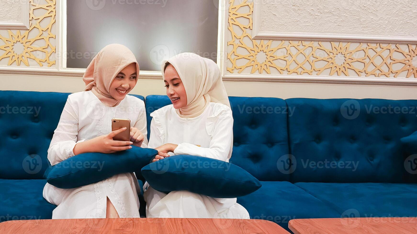 two young asian women candid smiles chatting something with islamic ornament calligraphy background in arabic nuance sitting on the sofa photo