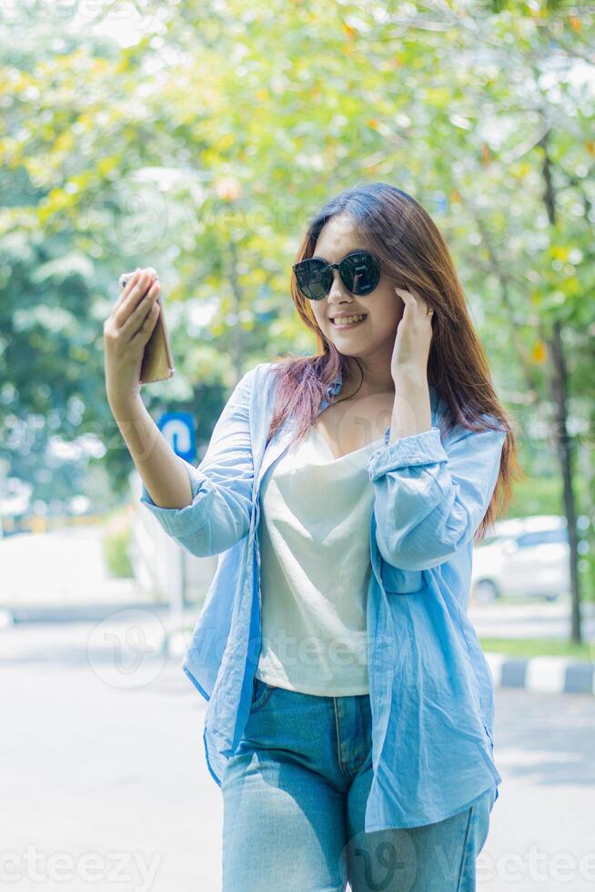 Casual indonesia girl in sunglasses smiling looking at camera carrying cellphone and earphones standing on outdoor park path. female students for the themes of education, fashion and lifestyle photo
