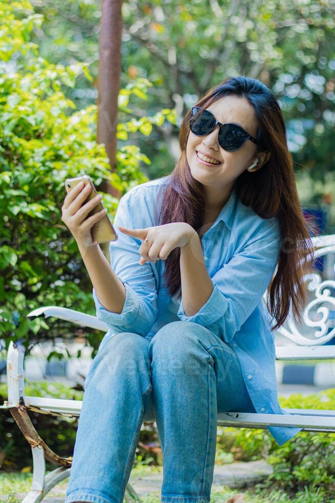 An indonesia mature woman in sunglasses looks at the camera cheerfully pointing at a cell phone gadget sitting in an outdoor park. female students for the themes of education, technology and lifestyle photo