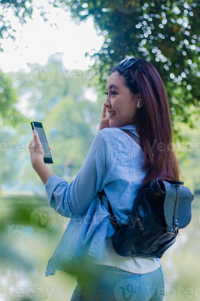 Beautiful Asian woman candid smile listening music via gadget smart phone in campus park on sunny summer day. female students for education, promotion and lifestyle concepts photo