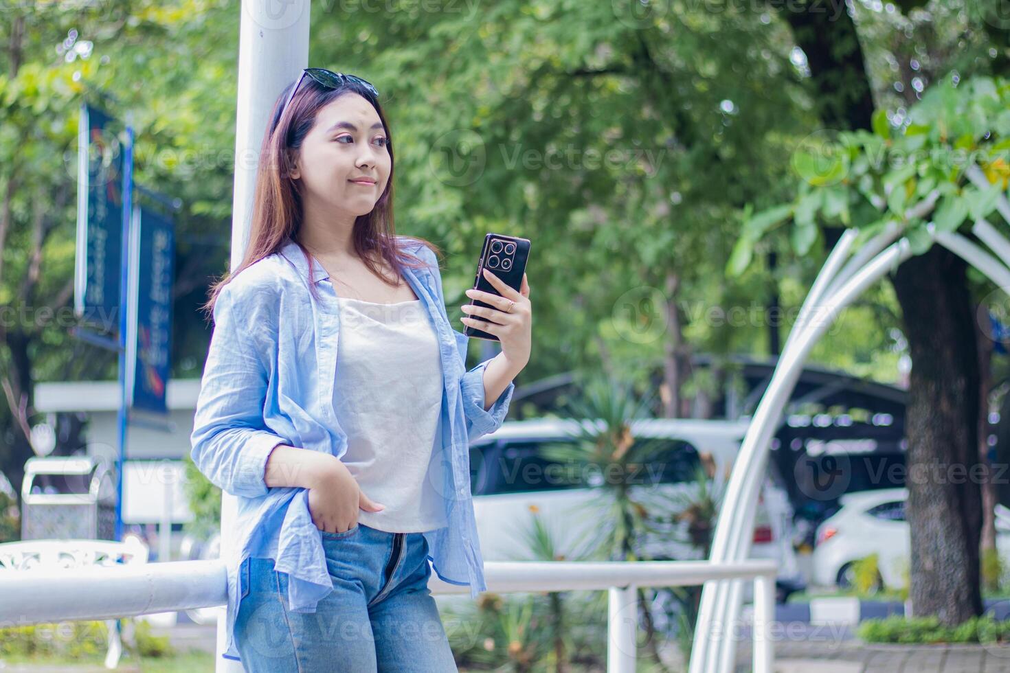 Candid beautiful indonesia woman leaning on the fence holding a smartphone gadget in the campus park in sunny weather. female student for education, promotion and lifestyle concept photo