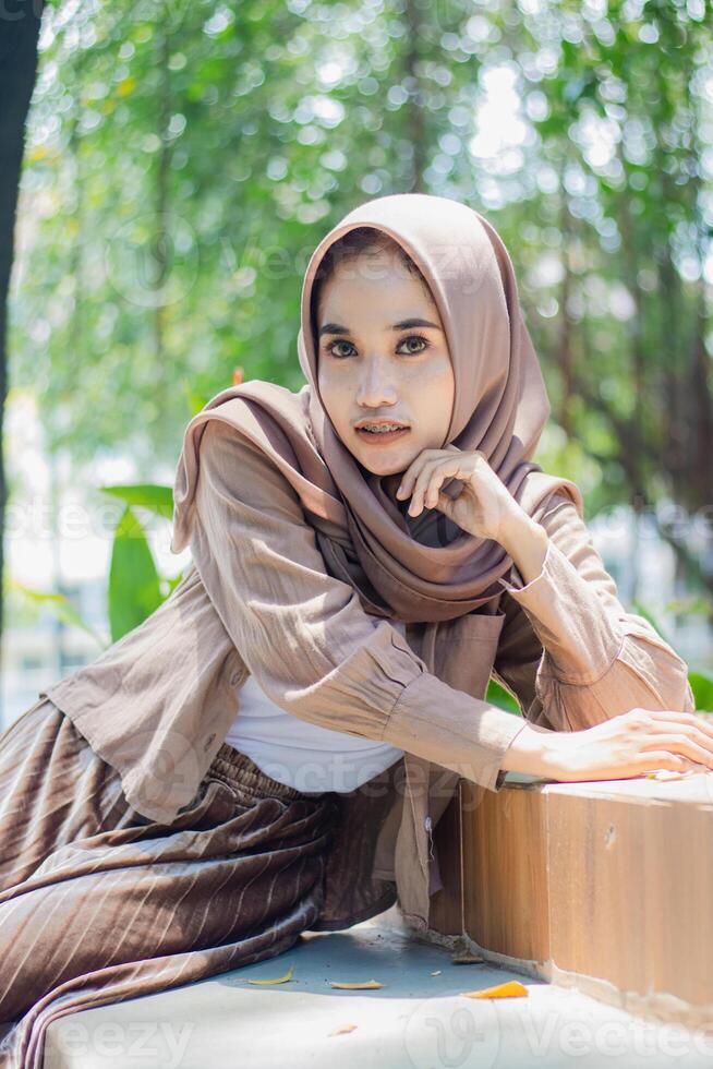 A beautiful elegant hijab Asian woman looks at the camera leaning against the ceramic and sitting on a chair in the outdoor campus park. student for the concept of human interest and lifestyle photo