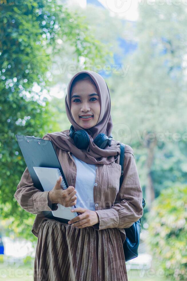 Young asian girl in hijab smiles gracefully at the camera carrying book and clipboard standing in outdoor park. female students for the themes of education, technology and advertising photo