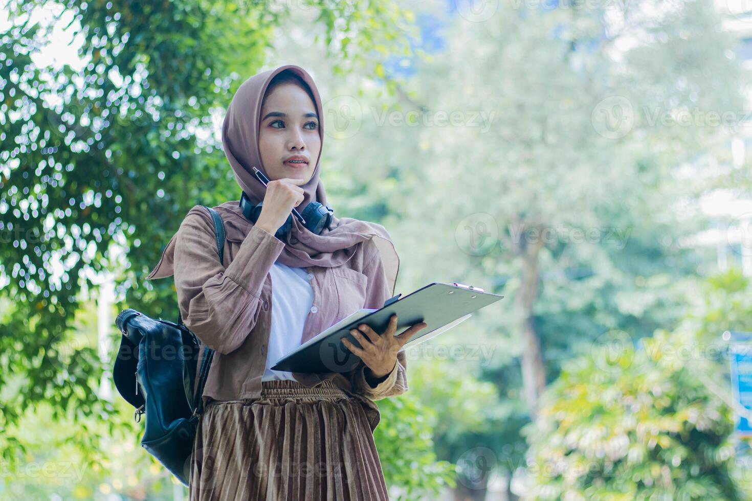 Young indonesia woman in hood thinking holding pen and carrying bag clipboard standing in outdoor park. female students for the themes of education, technology and advertising photo
